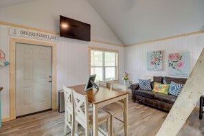 A room with a wooden dining table, white chairs, a brown leather sofa, a TV mounted on the wall, and a window with blinds. - Studio Cabin in Higden: Boating, Fishing & More! (Higden)