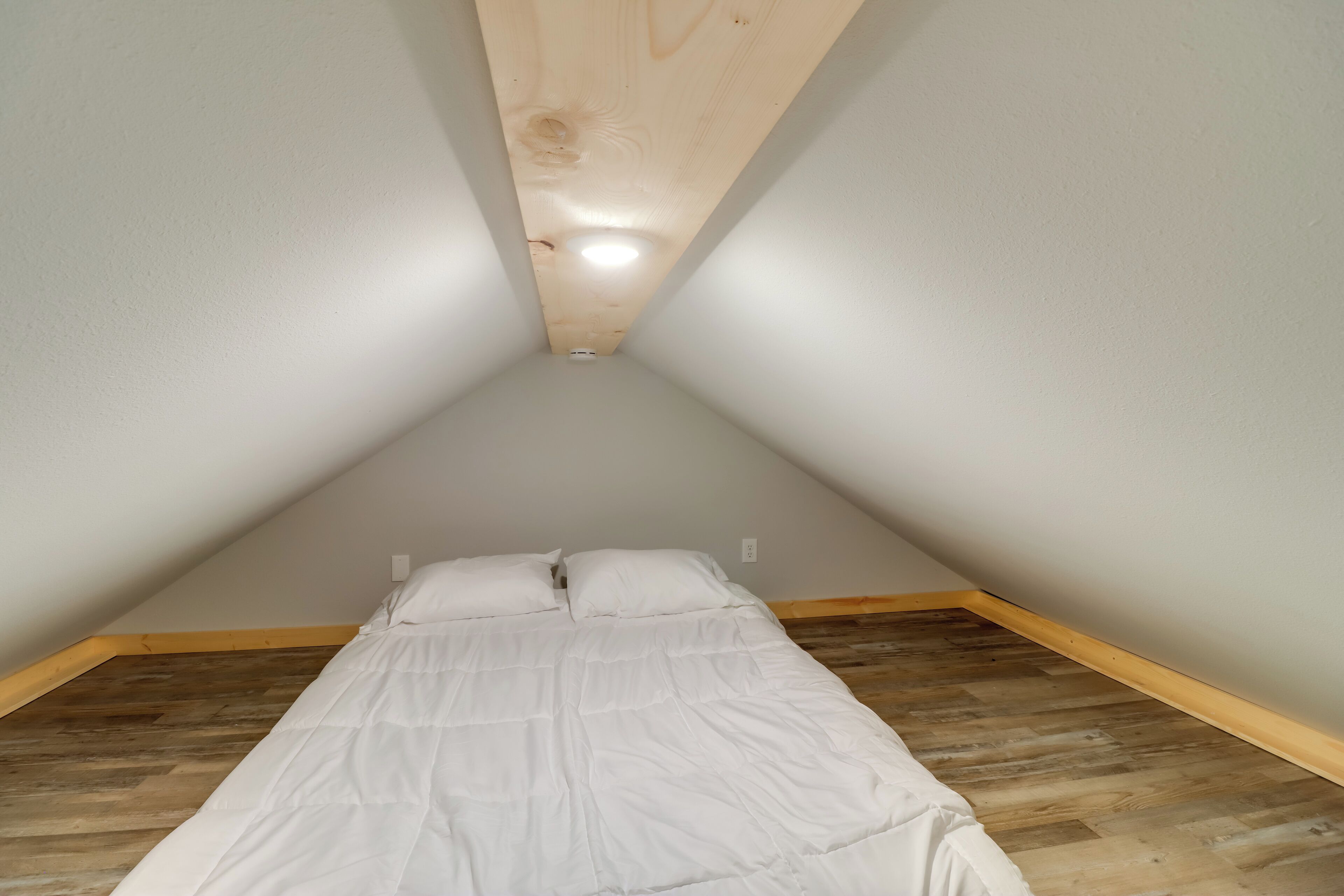 A small, triangular attic bedroom with a wooden floor, a white bed, and a sloped ceiling with a light fixture.