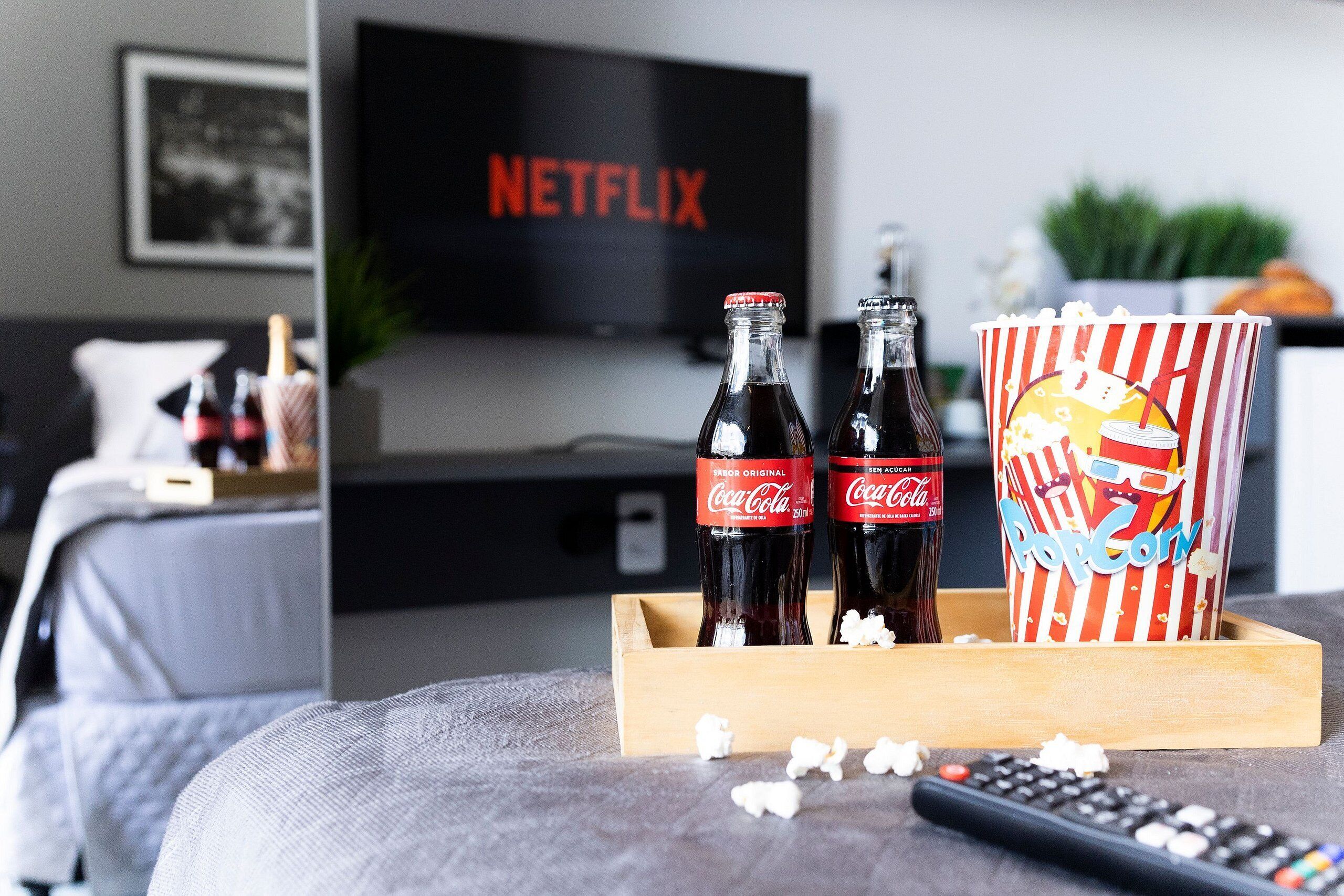 Ein gemütliches Wohnzimmer mit Fernseher, der 'NETFLIX' anzeigt, zwei Coca-Cola-Flaschen, einer Popcorndose und einer Fernbedienung auf einer grauen Couch.