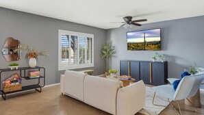A living room with a sofa, a TV mounted on the wall, a ceiling fan, and a window with blinds. - Pool Fire Pit  Ping-Pong (Tucson)