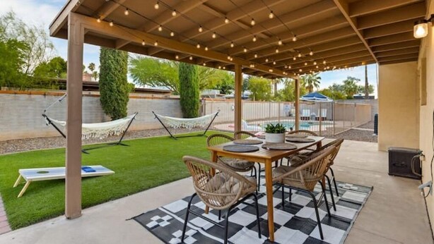 An outdoor patio area with a wooden pergola, string lights, a dining table with chairs, a hammock, and a small garden. - Pool Fire Pit  Ping-Pong (Tucson)