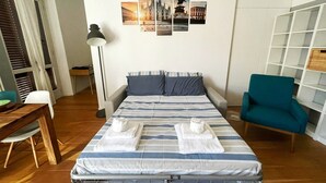Een net opgemaakt bed met gestreepte beddengoed en handdoeken, een bureau met stoel, een vloerlamp en een teal fauteuil.