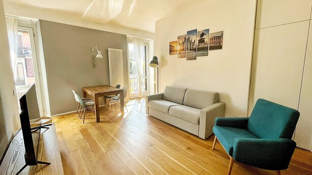 Een moderne woonkamer met houten vloer, een grijze bank, een blauwgroene fauteuil, een eettafel met stoelen en een tv-meubel.