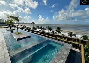 A pool with a view of the beach and ocean, surrounded by palm trees and a wooden deck. - LM UNITY CABO BRANCO (João Pessoa)