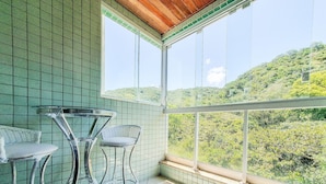 A balcony with a view of a green hillside, a glass table, and two wicker chairs.