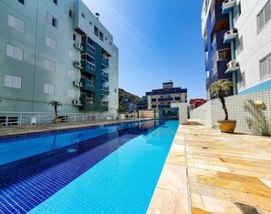 A swimming pool with tiled edges, surrounded by buildings with balconies and air conditioning units. - Residencial Ébano Marfim e Jequitibá (Ubatuba)