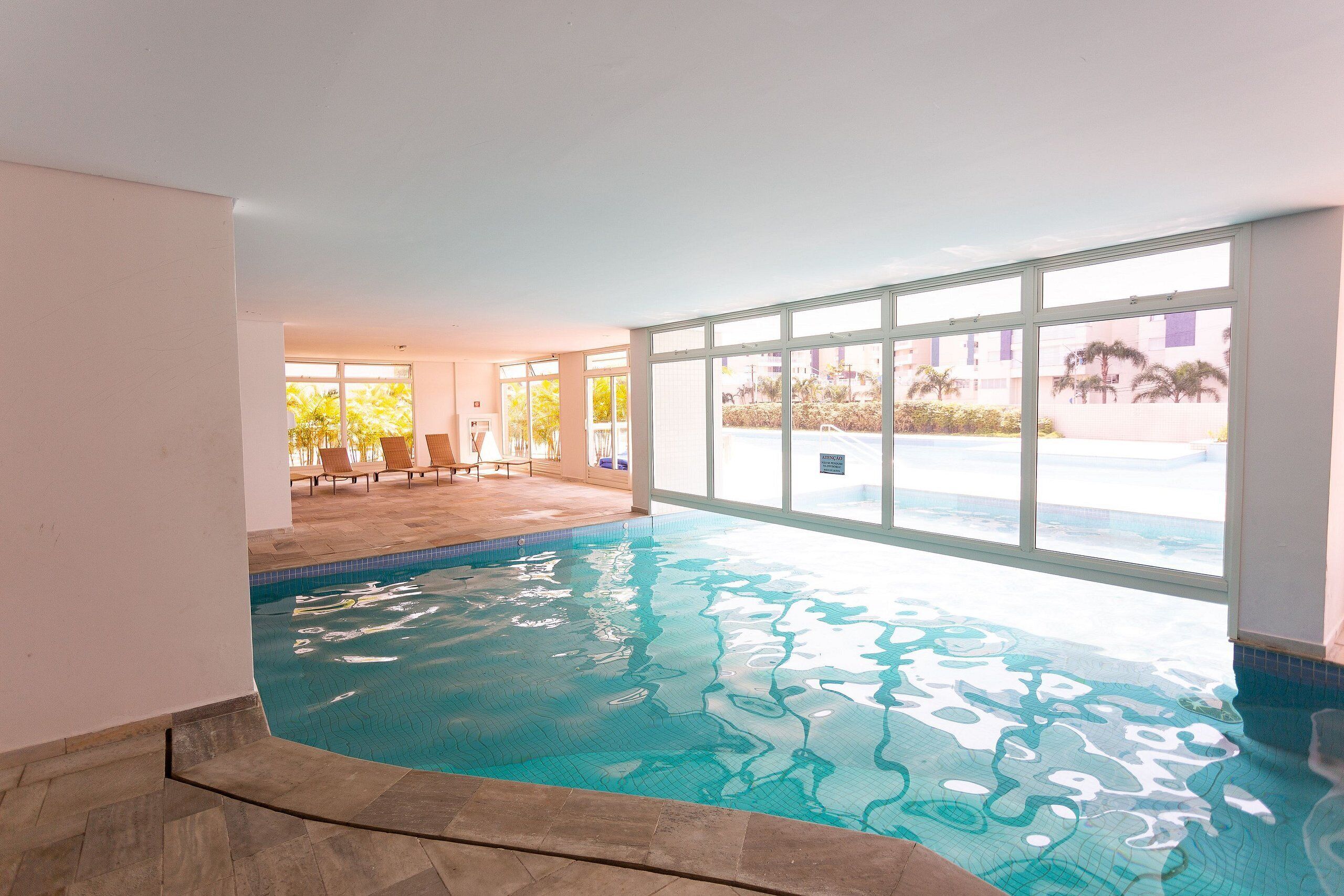An indoor swimming pool with clear water, a tiled floor, and a view of the outside through large windows.