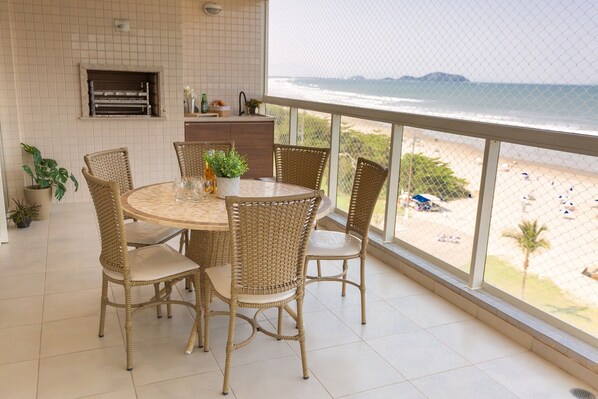 A balcony with a view of the beach, wicker chairs, a round table, and a tiled floor.