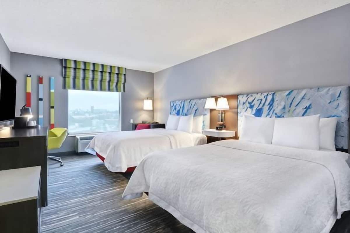 Ein Hotelzimmer mit zwei Betten, einem Schreibtisch, einem Stuhl, einem Fernseher und einem Fenster mit Blick auf die Stadt.