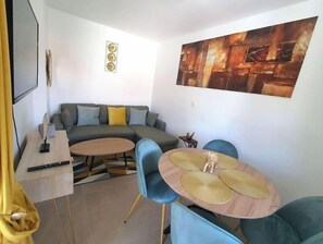 Una sala de estar moderna con un sofá modular, una mesa de comedor redonda con sillas azules, un televisor de pantalla plana montado en la pared y un cuadro abstracto grande sobre el sofá.