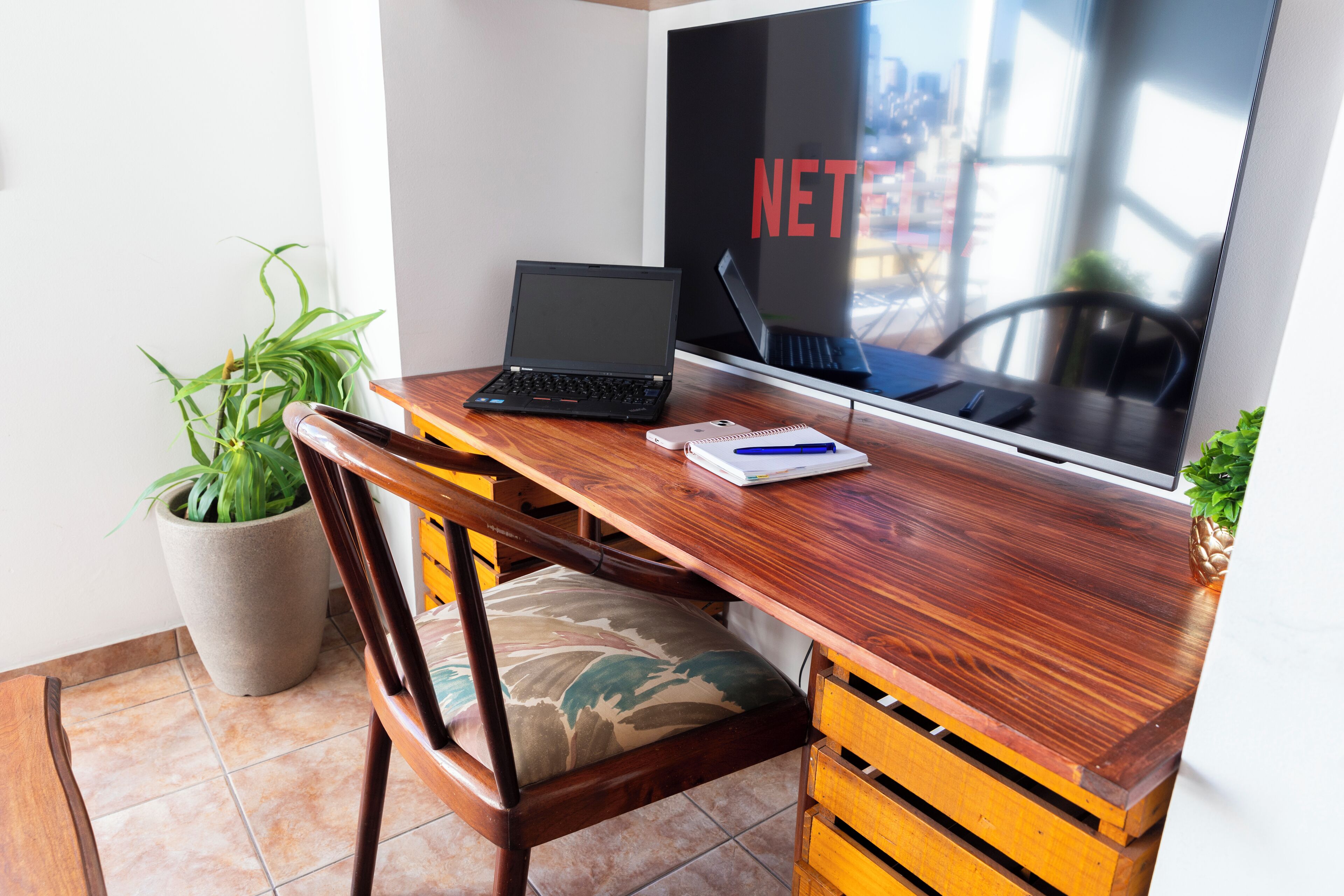 A wooden desk with a laptop, a notebook, and a pen. There is a potted plant and a chair.