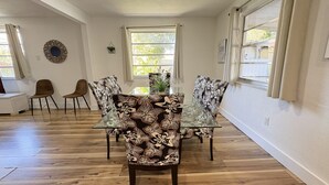 A dining area with a glass table surrounded by floral-patterned chairs, wooden flooring, and large windows with curtains. - Miami 3BR | Walk to LoanDepot Park | Patio (Miami)