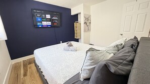A bedroom with a bed, a grey sofa, a mounted TV displaying streaming services, and a door with a painting.