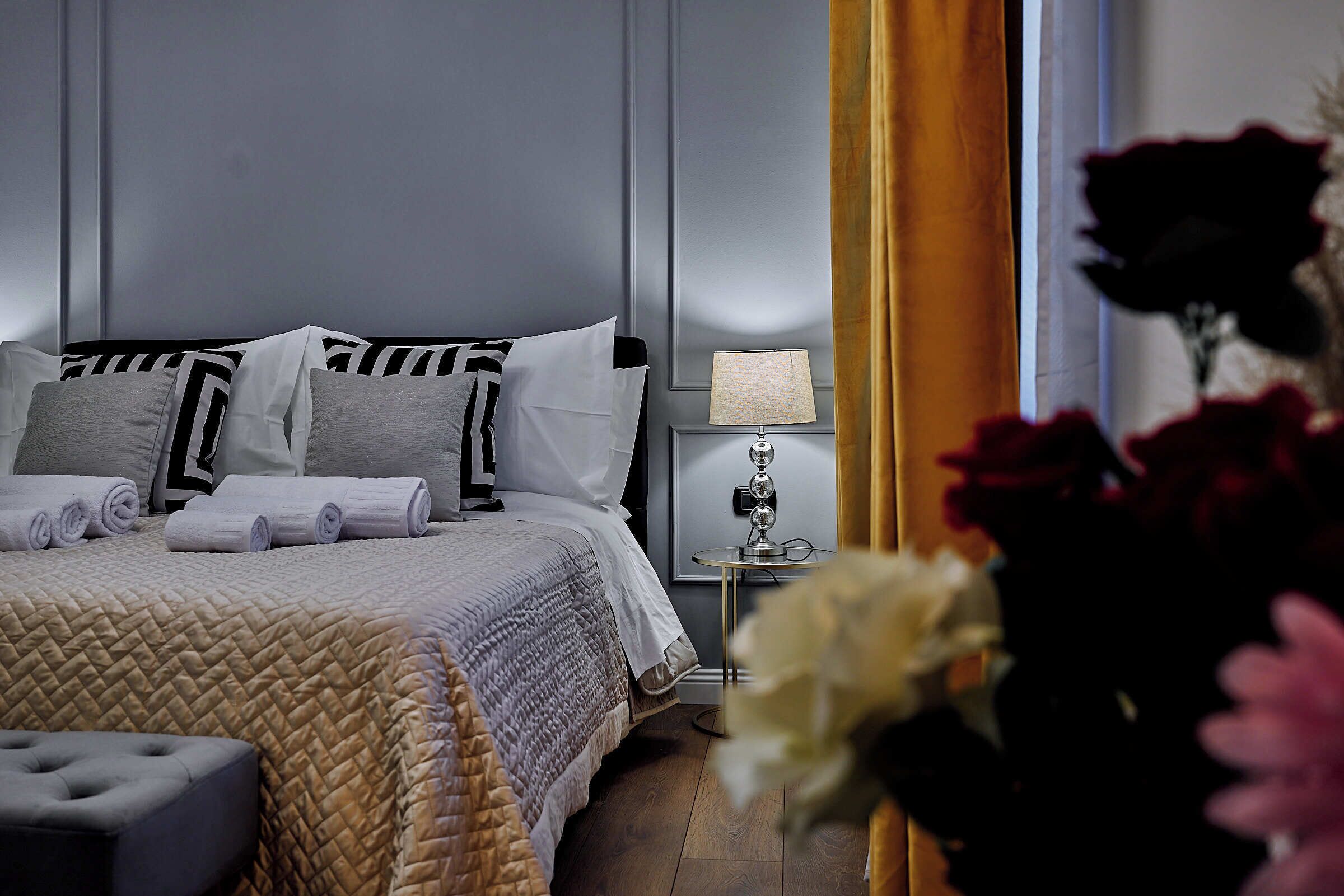 Uma cama bem arrumada com cobertura acolchoada, uma mesa de cabeceira com abajur e um vaso com flores.