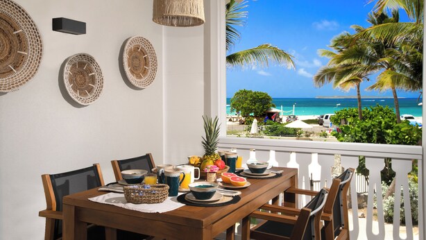 A dining area with a table set for breakfast, overlooking a beach with palm trees and the ocean.
