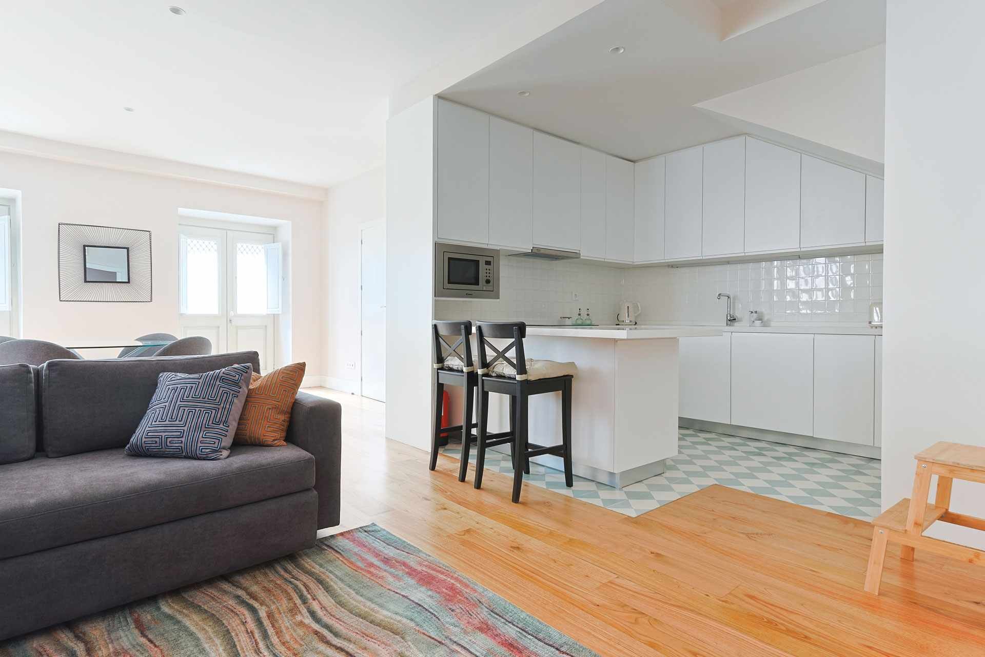 A modern apartment with a grey sofa, a colorful rug, a kitchen with white cabinets, and a small wooden table.