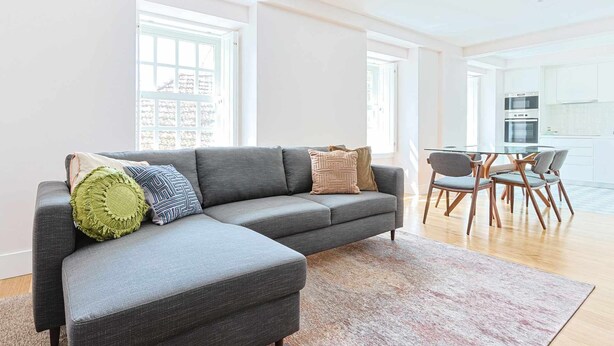 Ein modernes Wohnzimmer mit einem grauen Ecksofa, einem Glastisch mit vier Stühlen und einer Küche im Hintergrund.