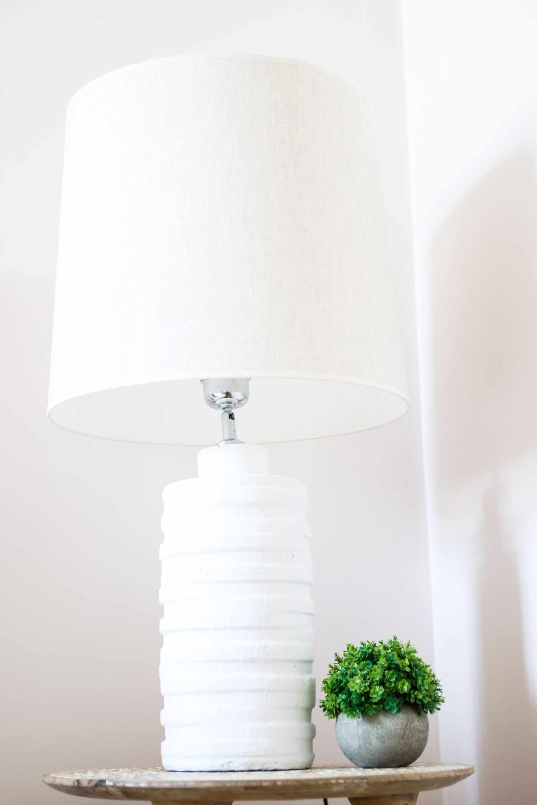 Une lampe blanche à motifs en relief, surmontée d’un grand abat-jour blanc, est posée sur une petite table ronde. Une petite plante en pot se trouve à côté de la lampe.