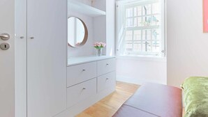 Ein Schlafzimmer mit einem weißen Kleiderschrank, einem runden Spiegel, einer kleinen Ablage mit einer Holzfigur, einem Fenster mit weißen Fensterläden und einem grünen Bett.