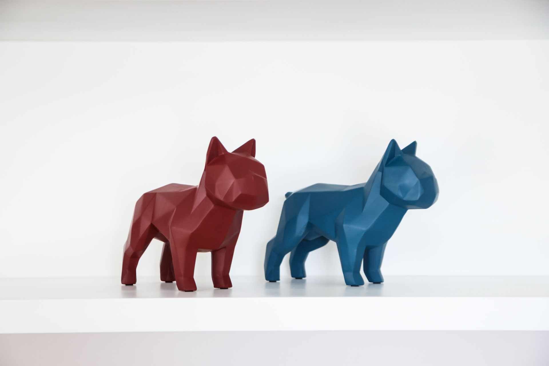 Deux sculptures de chats aux formes géométriques, l’une rouge et l’autre bleue, sont posées sur une surface blanche.
