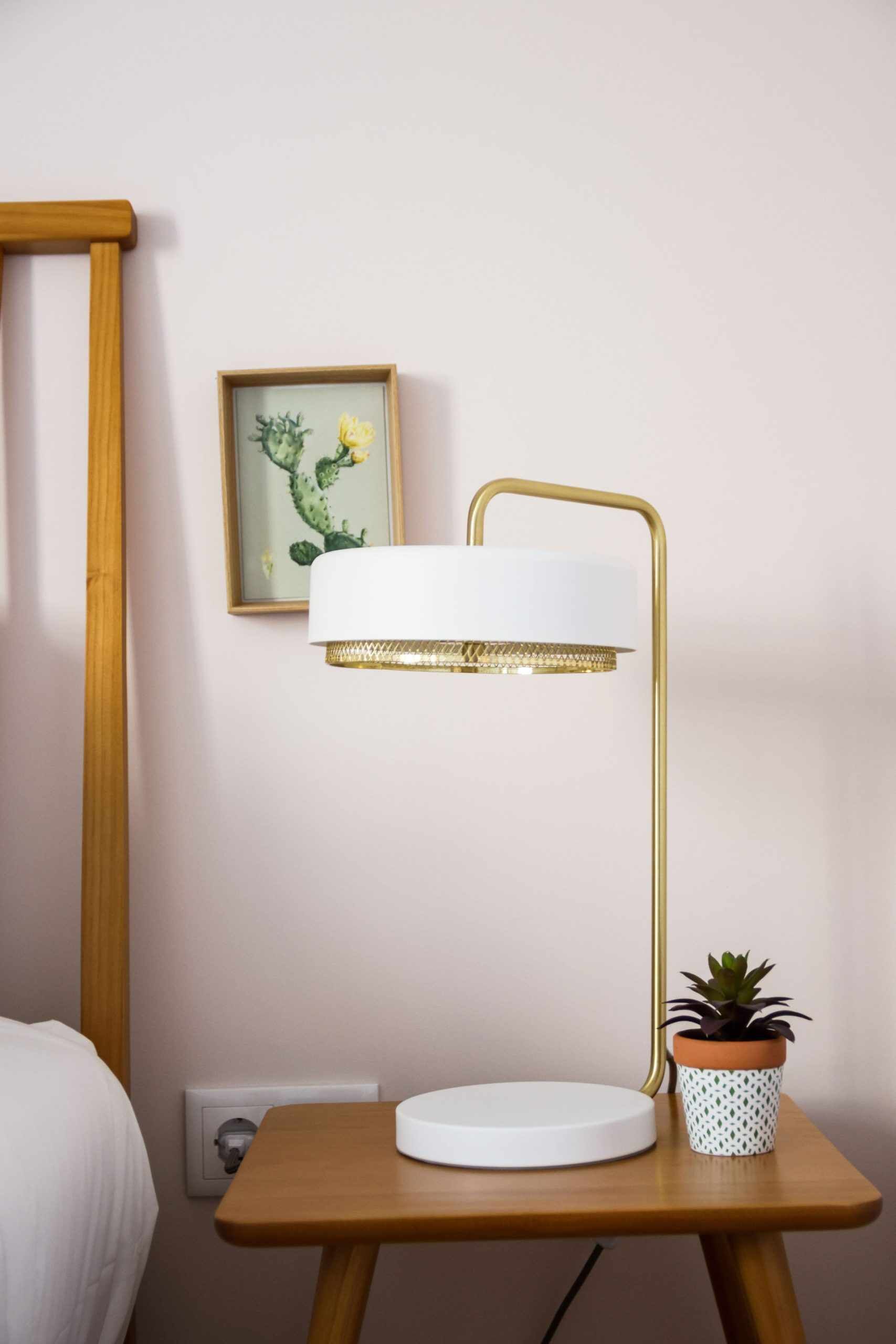 Ett litet bord vid sängen med en lampa, en liten krukväxt och en inramad tavla på väggen.