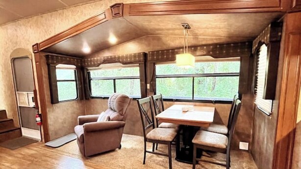Interior acogedor de una autocaravana con zona de comedor, una butaca mullida y una ventana que ofrece vistas a la vegetación.