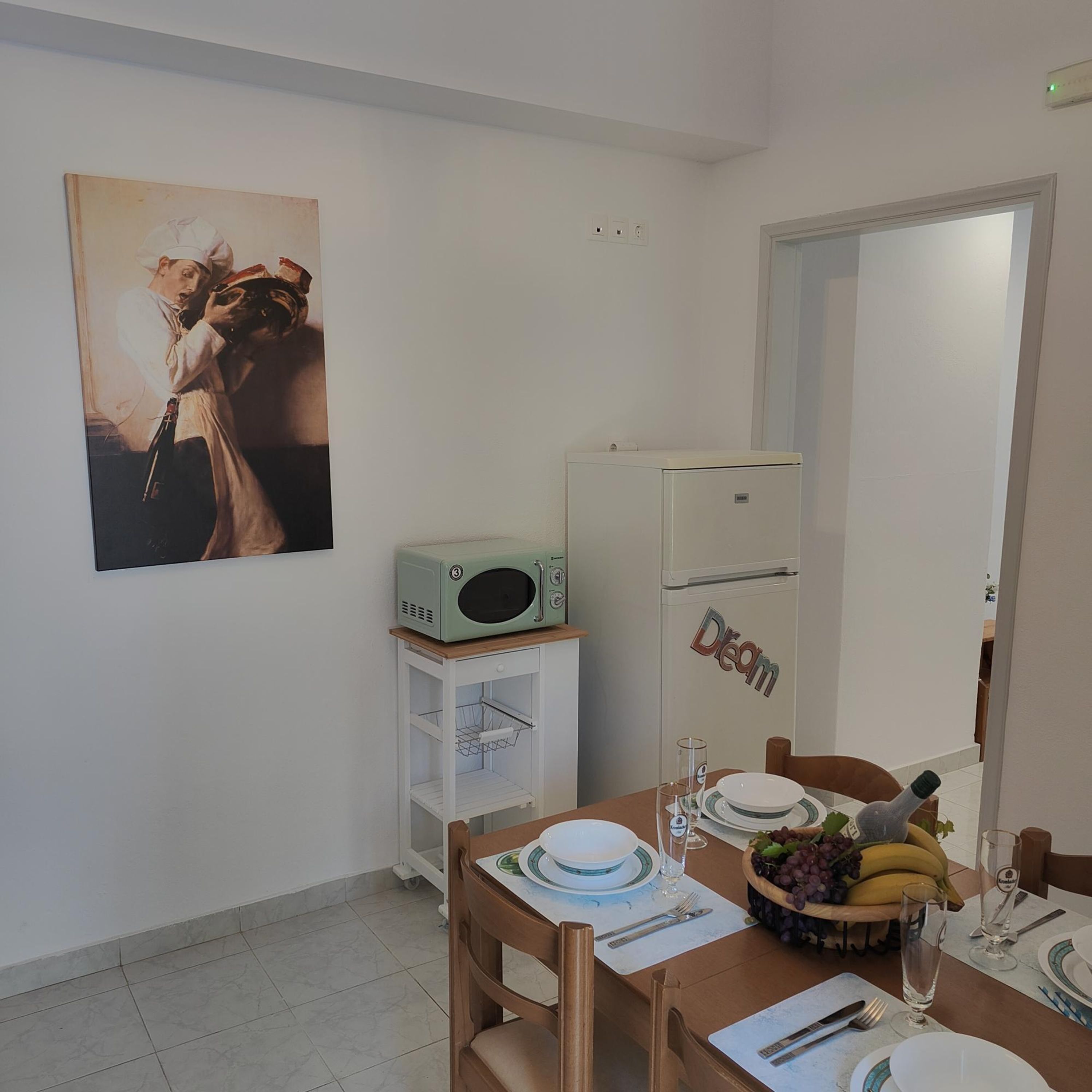 A dining area with a table set for two, a microwave, a refrigerator, and a painting on the wall.