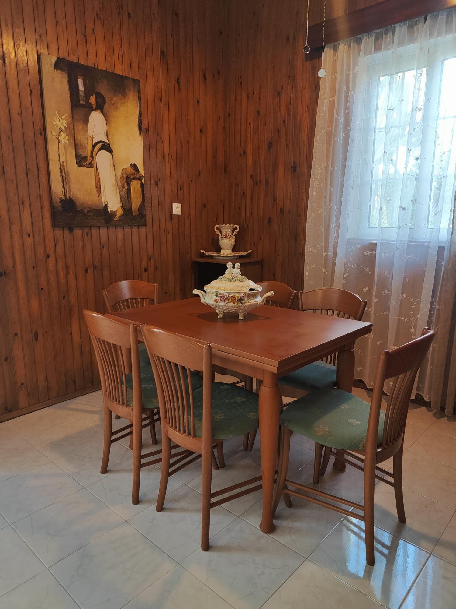 A dining area with a wooden table and chairs, a painting on the wall, and a window with curtains.