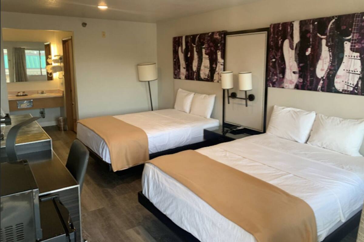 A hotel room with two beds, a desk, a chair, a mirror, and a painting of guitars on the wall.