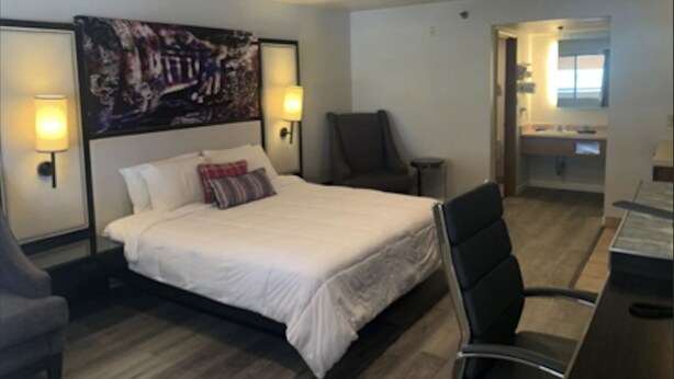 A hotel room with a bed, two wall-mounted lamps, a chair, a desk, and a painting on the wall. - GO2 Inn & Suites by Relianse  (El Paso)