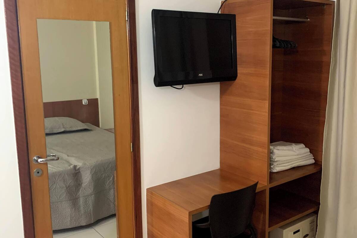 Ein Schlafzimmer mit einem Holztisch, einem Stuhl, einem an der Wand montierten Fernseher, einem Kleiderschrank und einem Bett mit grauer Bettdecke.
