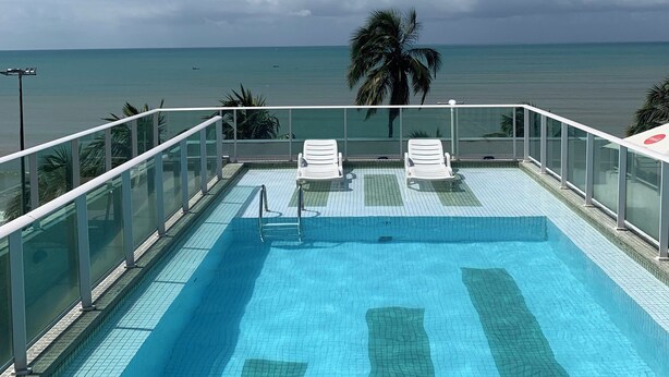 A clear blue pool with white lounge chairs and a glass railing overlooking the ocean. - Melhores Flats (João Pessoa)