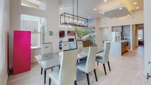 A modern dining area with a glass table and chairs, a red refrigerator, and a view of a landscaped area through large windows. - Balloon Fiesta: Rooftop Deck, Hot Tub, Sunsets (Albuquerque)