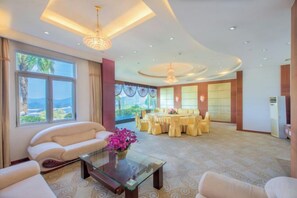 Ruime hotelkamer met een groot raam, een bank, een glazen salontafel en een eethoek met ronde tafels.