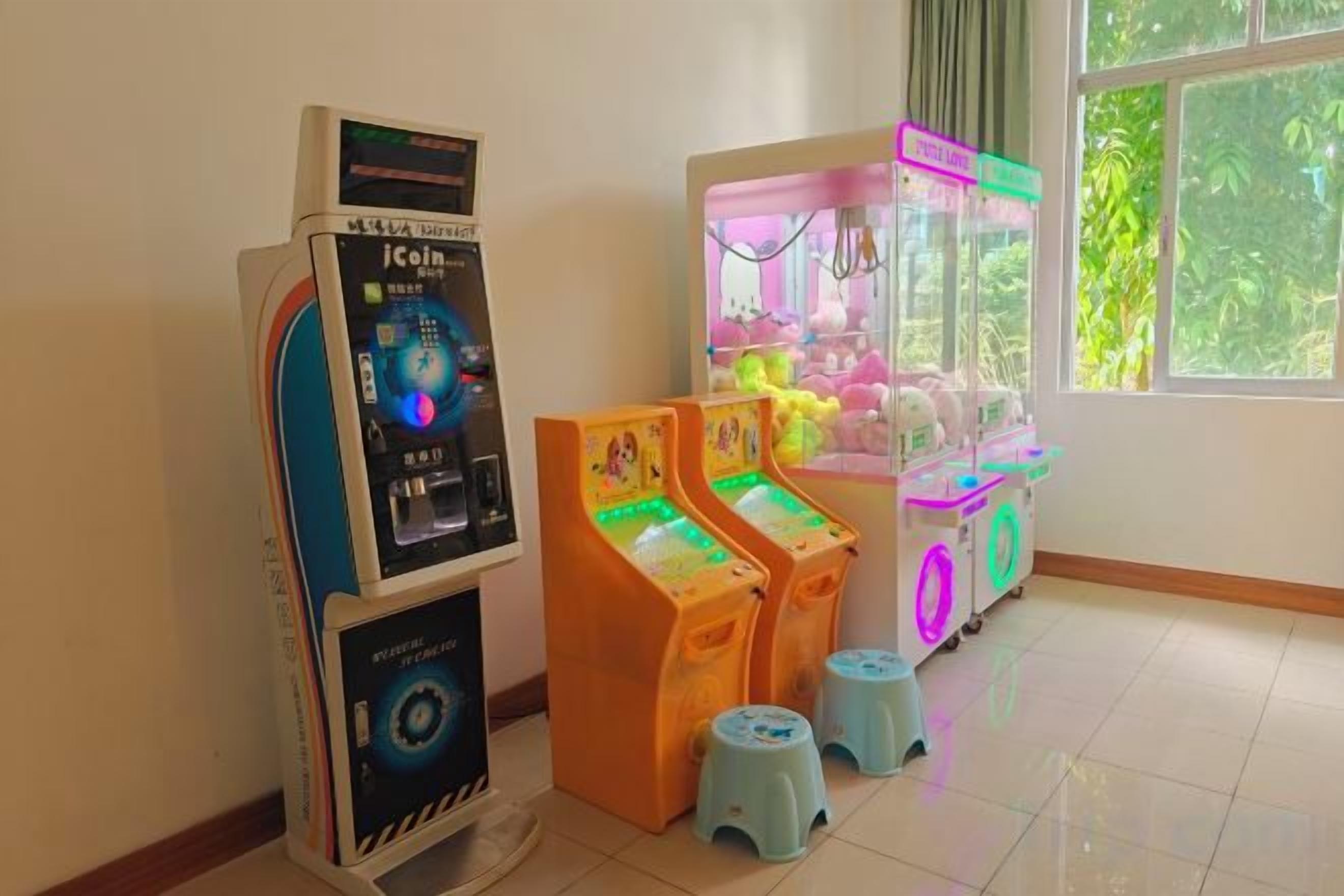 Una habitación con una máquina de juegos con monedas, dos máquinas de grúa y una ventana con vistas a zonas verdes.
