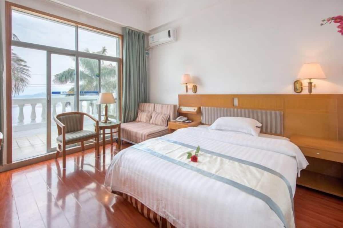 Une chambre d’hôtel avec un grand lit, une chaise, une petite table et une vue sur l’océan grâce à de grandes fenêtres.