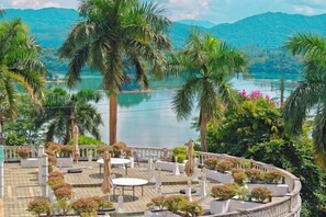 Une terrasse avec des sièges extérieurs, entourée de palmiers et offrant une vue sur un lac et des montagnes.