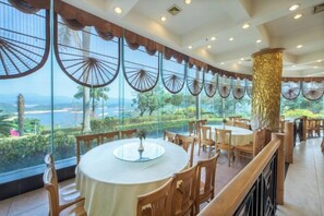 Un restaurant avec des tables et des chaises rondes, de grandes fenêtres ornées de motifs décoratifs en forme d’éventails, et une vue sur l’extérieur.