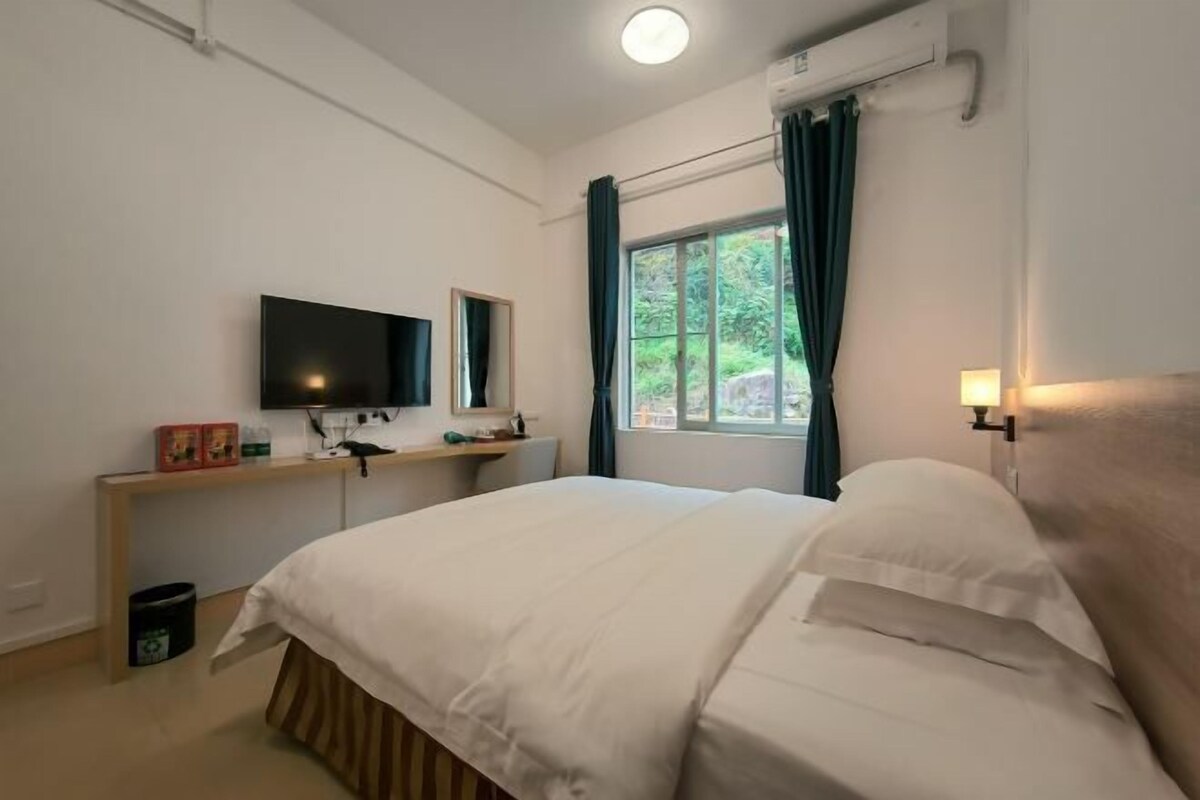A hotel room with a bed, a TV, a window with curtains, a bedside lamp, and a small table with items on it.