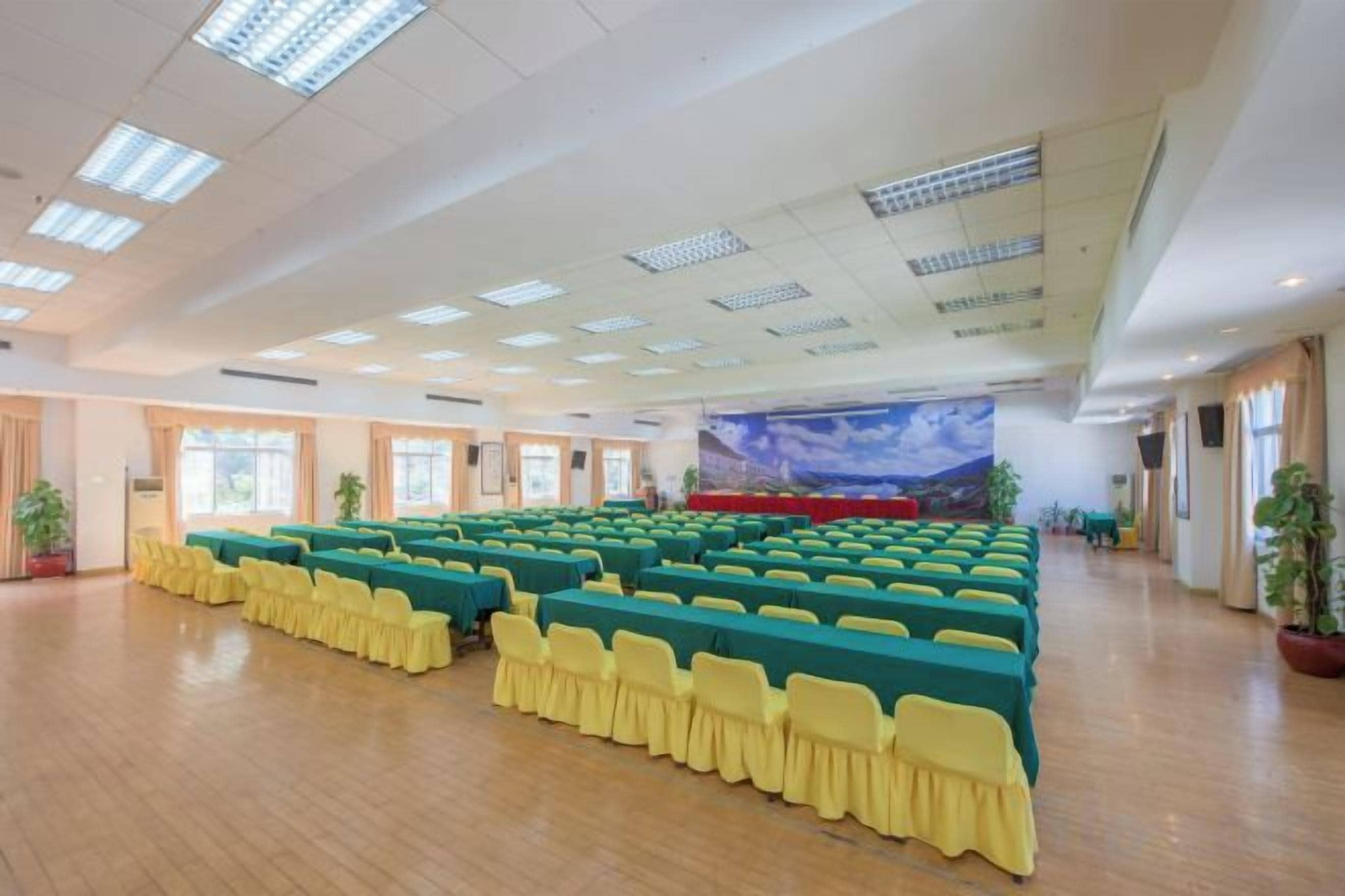 Un amplio salón de conferencias con filas de mesas y sillas, un escenario con fondo decorativo y plantas en macetas.