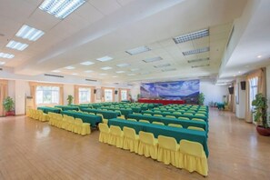 Een ruime congreszaal met rijen tafels en stoelen, een podium met decor en potplanten.