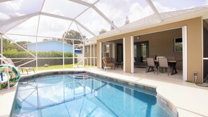 Une piscine dotée d’une structure en verre, un espace extérieur aménagé pour s’asseoir et une vue sur un bâtiment bleu en arrière-plan.
