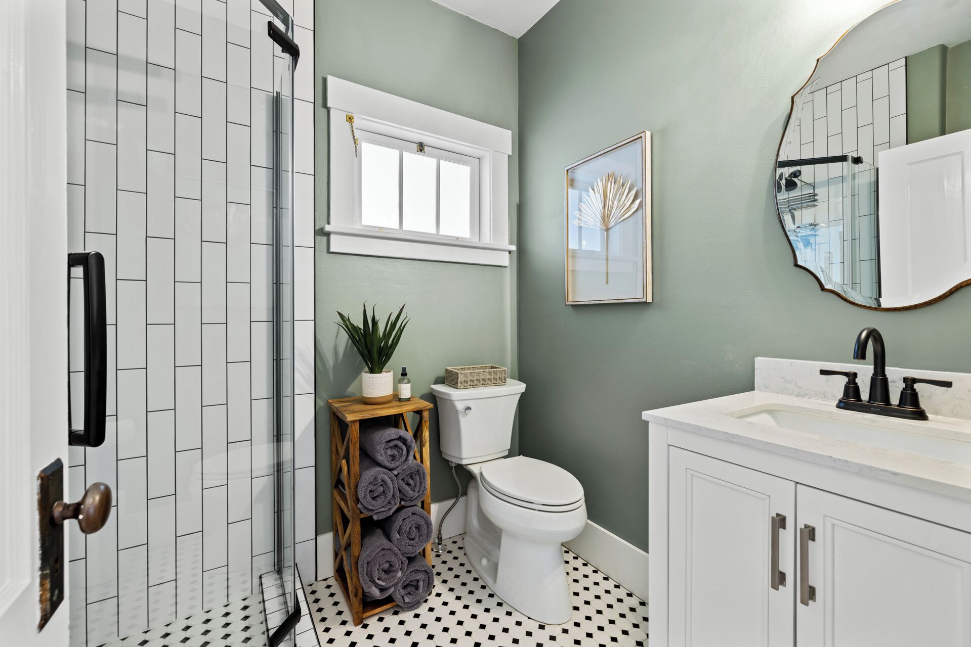 Un baño con un inodoro blanco, un lavamanos con grifo negro, un taburete de madera con toallas, una planta en maceta y una obra de arte enmarcada en la pared.