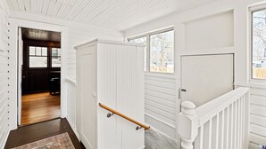 Een gang met witte wanden en een houten vloer, die leidt naar een kamer met een donker houten deur en een raam.