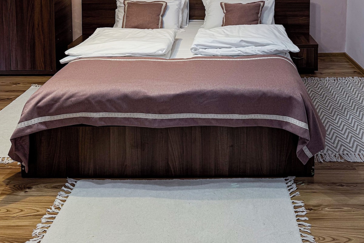 Un lit double bien fait, avec une tête de lit marron et du linge de lit blanc, une armoire en bois sur le côté gauche et un ventilateur de plafond avec des lampes.