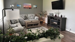 Un salon moderne avec un canapé gris, une table basse en bois, une lampe sur pied, un téléviseur fixé au mur et plusieurs tableaux accrochés au mur.