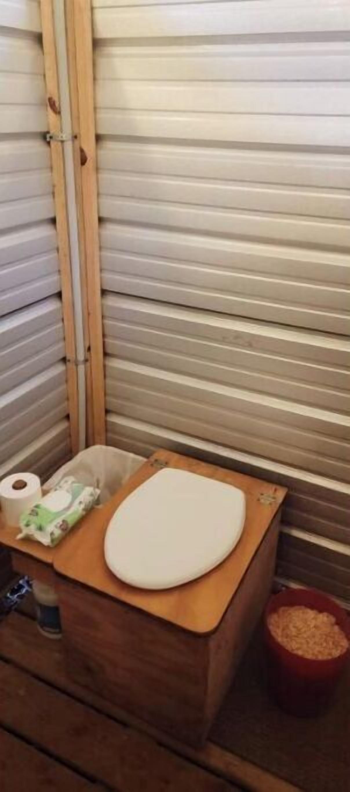 A small, enclosed space with a wooden bench, a toilet, and a trash bin.