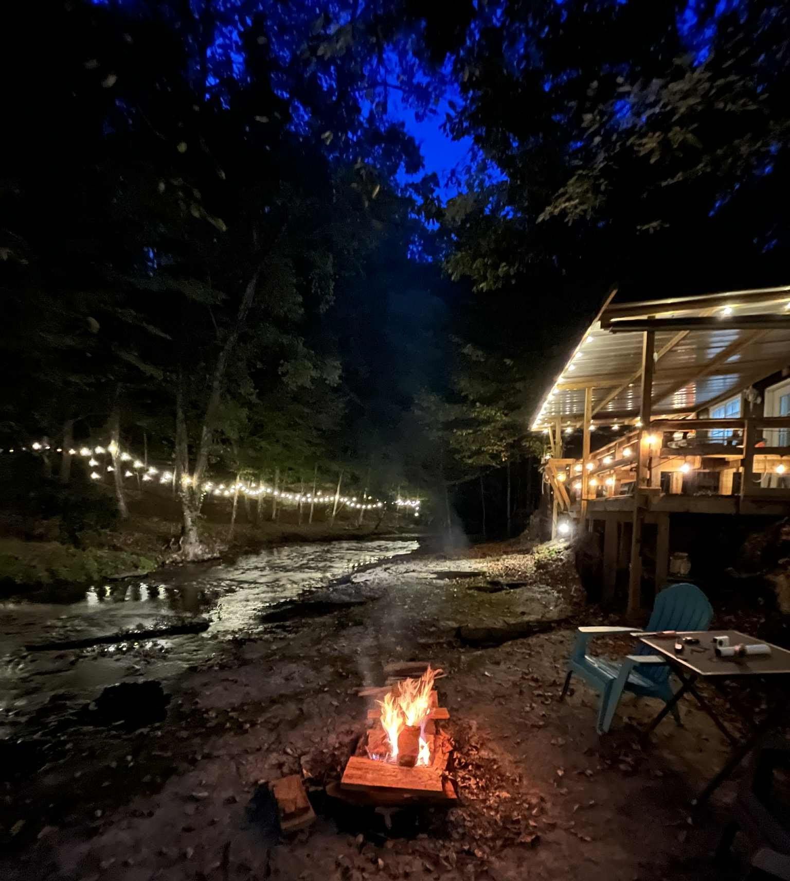 A riverside outdoor seating area with a fire pit, surrounded by trees and a building with illuminated windows.