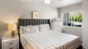 Een slaapkamer met een bed, nachtkastje, lamp, plafondventilator, raam met uitzicht en een lijst met foto aan de muur.