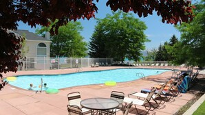 Une piscine entourée d’arbres et d’une clôture, avec des chaises longues et des tables à proximité.